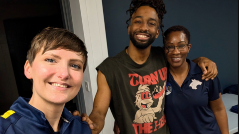 Two students are pictured with a male dancer backstage at the show in Birmingham. The students have navy tops on with a Wolf emblem on. One has short brown hair the second cropped hair and glasses on. The male has a dark grey T-shirt on which says "Turn up the heat". All are smiling at the camera.
