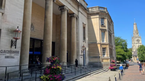 Street view in Gloucestershire, England, featuring Shire Hall, home to Gloucestershire County Council. The entrance is framed by tall columns and hanging flower baskets, with pedestrians passing by. In the distance, a historic church spire rises above the treetops.