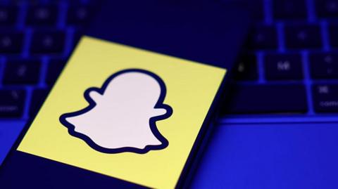 A phone displaying a Snapchat logo on top of a laptop in a darkened room.