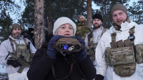 A woman dressed in a dark coat, navy gloves and cream hat stands looking above a pair of binoculars she is holding, She stands surrounded by several soldiers dressed in snow-white uniform, armed with camo belts and guns. The group appear to be in a snowy forest. 