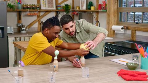2 men performing an experiment using bottle with balloons on top of them. They are sat at a workbench and wearing a yellow t shirt and a green shirt.
