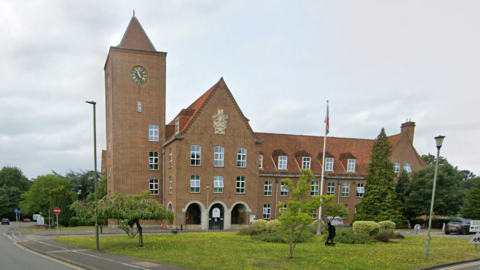 The red brick headquarters of Spelthorne Borough Council, a red-roofed building with slanted rooves, a clock tower and a green space and roads in front of it. A flag flies from a flagpole on the green space.
