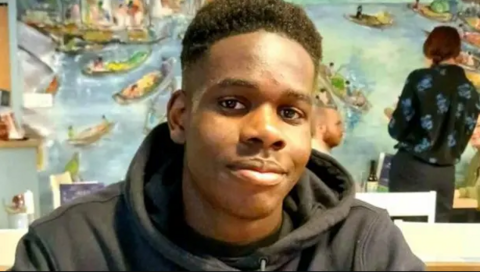 Teenager Shayne Hambakachere in a black hoodie smiles at the camera with his mouth closed. He is sat at a table in a restaurant, with a few tables behind him. He has short hair with tight curls.