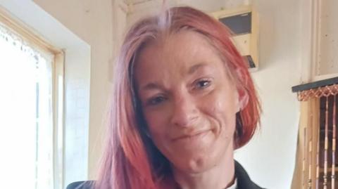 Reanne is smiling to the camera with her head tilted to her right. She has faded, dyed red hair that is swept down the right hand side of her face.