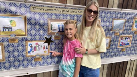 Ellie and her mum stand and point at Ellie's artwork printed on the wall beside her. They both have blonde hair and are smiling. Her mum wears black sunglasses and Ellie is wearing a multicoloured tie-dye t-shirt.