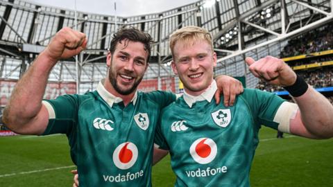 Caelan Doris and Jamie Osborne in a celebratory pose, arm in arm, with each man raising a clenched fist and smiling, while wearing Ireland's green shirt
