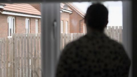 Anonymous image of a mother we are calling Caroline. She has dark hair. Her image is blurred to protect her identity. She is looking though a window into a back garden with a wooden fence and a row of newish houses in the background.