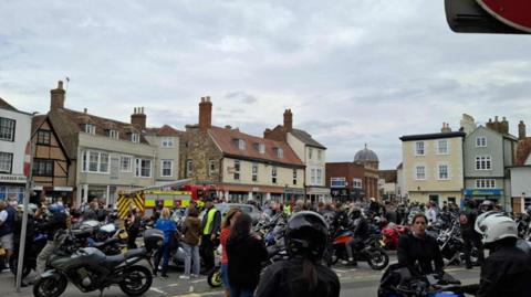A crowd of people, some wearing helmets, and a number of motorbikes among the crowd. A fire engine is among the crowd and there are shop buildings beyond.