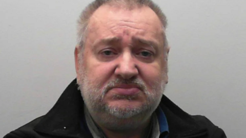 Ian Mark Wrightson has short grey hair and a short grey stubble. He is wearing a black jacket, against a grey background.
