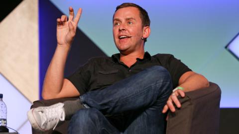 Scott Mills gestures as he speaks onstage wihle wearing a polo shirt, jeans and trainers, sitting down against a colourful backdrop, at the Edinburgh Fringe Festival in August 2014.