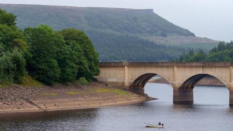 A reservoir with a single fisherman fishing from a boat in the middle. The level is very low, exposing a lot of bank on the left hand side. There's a bridge going across it, and hills and trees in the background.