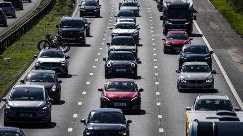 Several cars drive on a motorway. All three lanes are jam-packed. The cars are driving towards the camera. The shot is taken from above.