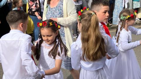 Five children dressed in white with floral head dresses dance through the streets of Helston, watched by a crowd of happy people 