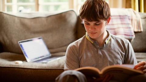 GCSE History student sitting in front of his sofa at home with a text book, listening to a BBC Bitesize revision podcast on ear buds.