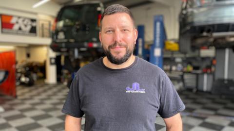 James Larbalestier has short brown hair with well groomed dark facial hair. He is wearing a navy blue T-shirt with his work logo emblazoned on it.