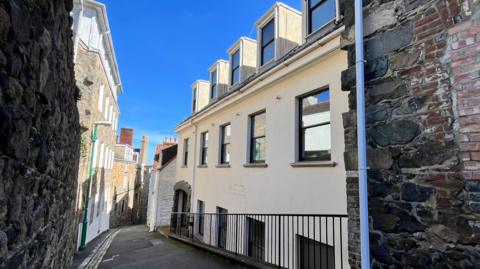 A three-storey office building on a lane in St Peter Port. The image is taken at the top of a hill, looking down. The building is to the right of the image. It is painted cream. The words 'islands insurance' are faintly visible on the wall. The sky is blue. 