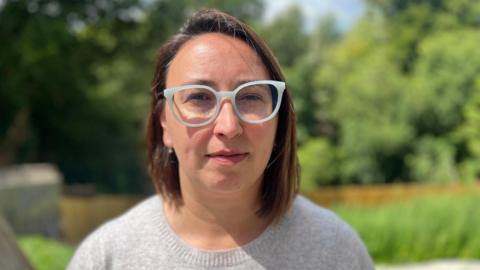 Dr Ralitsa Hiteva looks at the camera. She has short brown hair and is wearing a grey jumper. She also has glasses with white frames and is standing in front of a green background.