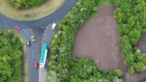 An aerial shot of a large patch of land off a busy roundabout. The patch of land is surrounded by trees. The land has been torn up and there is just brown soil visible now. 