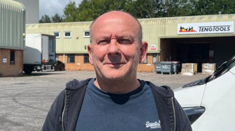 A bald headed man wearing a blue T-shirt and zip up top smiling into the camera. He is standing in an industrial estate with a van and tool company in the background.