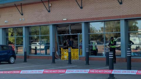 A modern glass and brick shop frontage with one section smashed inwards and a yellow barrier covering the gap and fire service warning tape between bollards