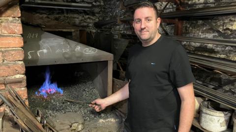 Simon Hall stands smiling at the camera while his right hand holds a poker into the blue and red flames of his forge.