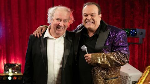 Shaun Williamson reprises his iconic role of Barry Evans. He is wearing a glittery suit jacket, and holding a microphone.