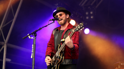 Gaz Coombes from Supergrass on stage with a black guitar, red jacket, sunglasses and black hat with purple and yellow lights behind him