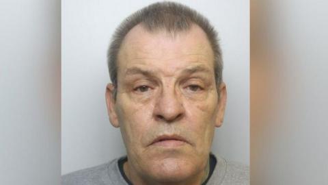 Sydney Burdon looking serious at the camera for his mug shot picture. He has a receding hairline and brown hair, wearing a grey top on. 