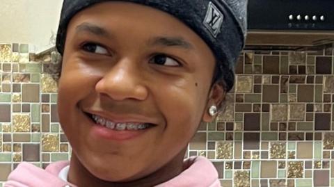 A close up picture of Leonardo who has braces and is smiling, looking away from the camera. He is wearing a Louis Vuitton grey beanie hat and has a large stud in his right ear. 