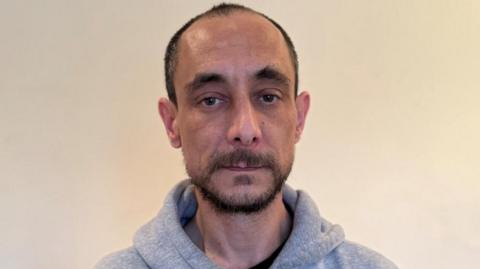 Steve looks at the camera and wears a grey hooded jumper. He has a dark beard, brown eyes and is partly bald. The background is blank.