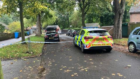 A police car parked in the middle of a road with blue police tape strung across the crime scene. The road is tree-lined, with fallen leaves on the floor and cars parked down both sides.