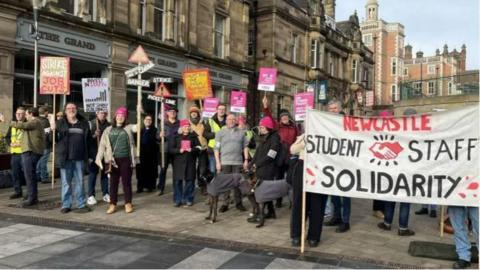 Men and women line a town square pavement, holding up posters, banners and placards that read 'strike against job cuts' and 'strike picket line'. There is a row of buildings behind them. 