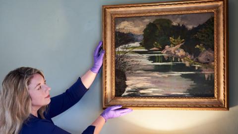 A woman with blonde hair, with purple latex gloves on and wearing a blue top, is holding a gold-framed landscape painting on a white wall