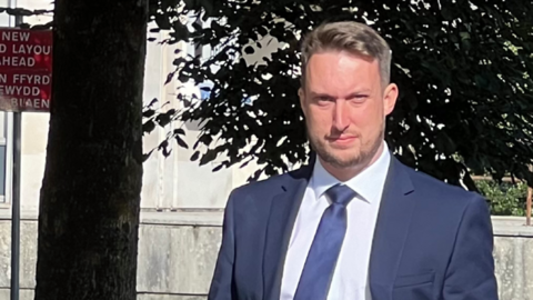 Huw Orphan, wearing a blue suit, white shirt and tie and carrying a bag, walks near Cardiff Crown Court and looks at the camera