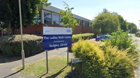 A blue and white sign with the Dudley Council and NHS logos reading "The Ladies Walk Centre Encompassing Sedgley Library". It is on a patch of grass with a pavement around it and a small street sign partially covered by shrubs. There are more shrubs and trees in the background and a red brick building with windows.
