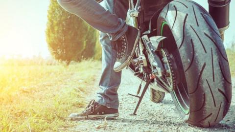 A biker's feet next to the wheels of a motorbike while it stands on its kickstand. The feet wear converse trainers with white toe tips and the rest of the footwear is dark. They stand on a gravel path next to a grassy field. The bike's wheels have dust on them.