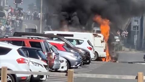 Flames are erupting from a white van and plumes of black smoke are going up into the sky. The van is parked in a row of other cars and people can be seen in the background leaving the scene.