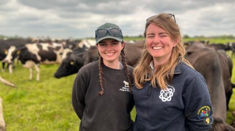 Emily and Georgie Paul stand in front of their cattle, on a big grassy field. They are both smiling, and have sun on their faces