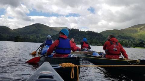 Four people, viewed from behind, canoeing on a lake. Green hills can be seen beyond the water.