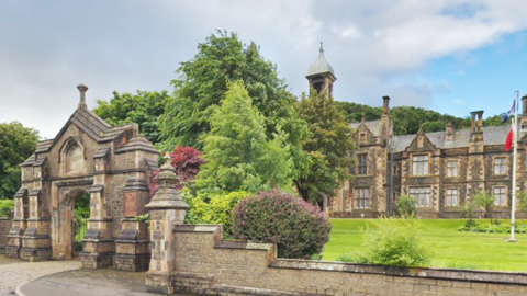 A large care home building, with a gated entrance and several large trees surrounding it