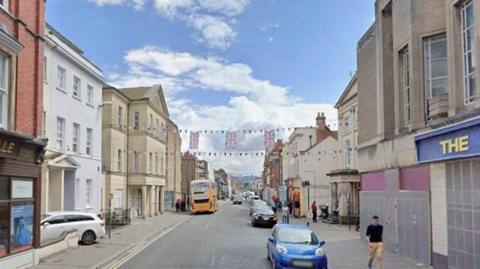 A shopping street in Gloucester city centre. There are several boarded up shops and cars parked along the road and the buildings are in different neutral colours and styles. There is Union flag bunting strung across the street.