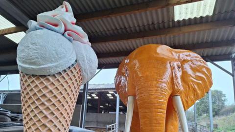 A giant ice cream cone statue next to a large orange elephant statue stood in a barn on a Devon farm