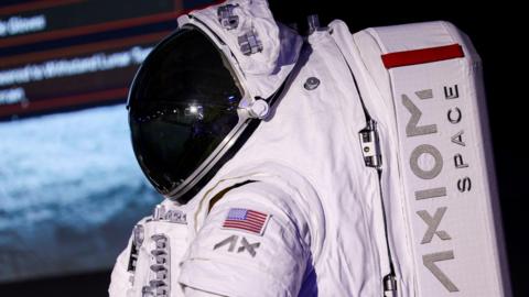 A white space suit complete with helmet and black visor is seen. It has a US flag on the arm and it says Axiom Space on the backpack. 