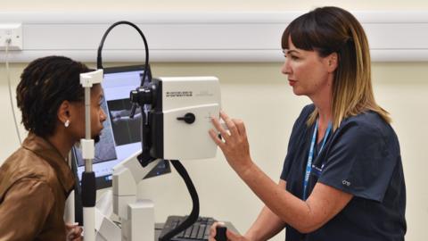 A nurse giving a woman an eye screening test. Both are seen side on. The nurse, on the right, has brown hair, and is wearing a blue uniform. One hand is adjusting a dial on a machine, similar to a large camera. The woman to the left is looking into the machine with her chin resting on a frame. She has dark braided hair and is wearing a brown leather jacked. A screen can be seen to the side of but the images it is displaying cannot be made out.