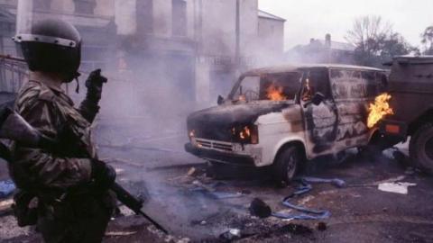 A British Army soldier stands with his back turned near a burning van as debris lies around him on a Belfast street. 