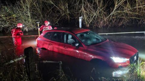 A red car stuck in water. The headlights are on and there are firefighters standing around the car in water 