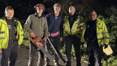 Five people, stood in a line, facing the camera. There are three police officers all in uniform, and high-vis jackets, and two young men wearing dark clothing. One is holding a chainsaw and the other a leaf blower. Foliage is behind them. 