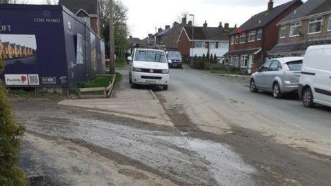 The entrance to the housing development, with what looks like mud and water going from the edge of the road into the entrance. There are boards at the site entrance saying Cube 鶹Լs and including some computer generated images of some of the properties. There are houses over the road, with some cars parked and a van parked half on the pavement and half on the road.