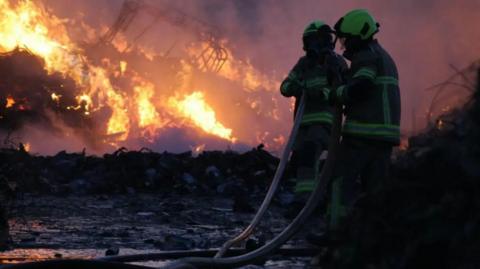 The image shows a nighttime scene involving two firefighters. They are dressed in full protective gear, including helmets and uniforms with reflective stripes. The firefighters are working together to manage a fire hose. The fire is intense, with bright flames and thick smoke rising from what appears to be a pile of waste. The flames provide the main source of light in the otherwise dark setting.