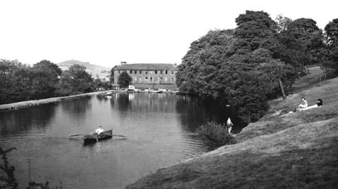 Roughlee Boating Lake (c.1955) showing two people in a rowing boat in the water with two women lying on the grass on the riverbank and a male standing near the lake in swimming trunks